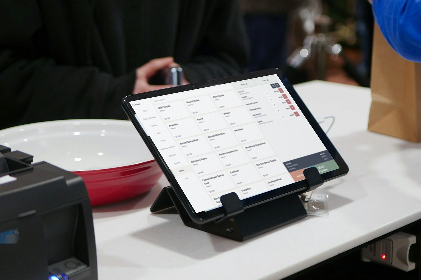 POS tablet in a stand on a club bar counter, showing a spirits menu and an open order, beside a receipt printer